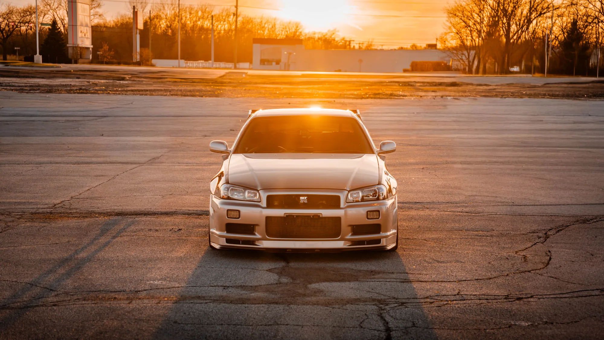 Silver Nissan Skyline R34 GT-R backlit by a low golden sun in an empty lot